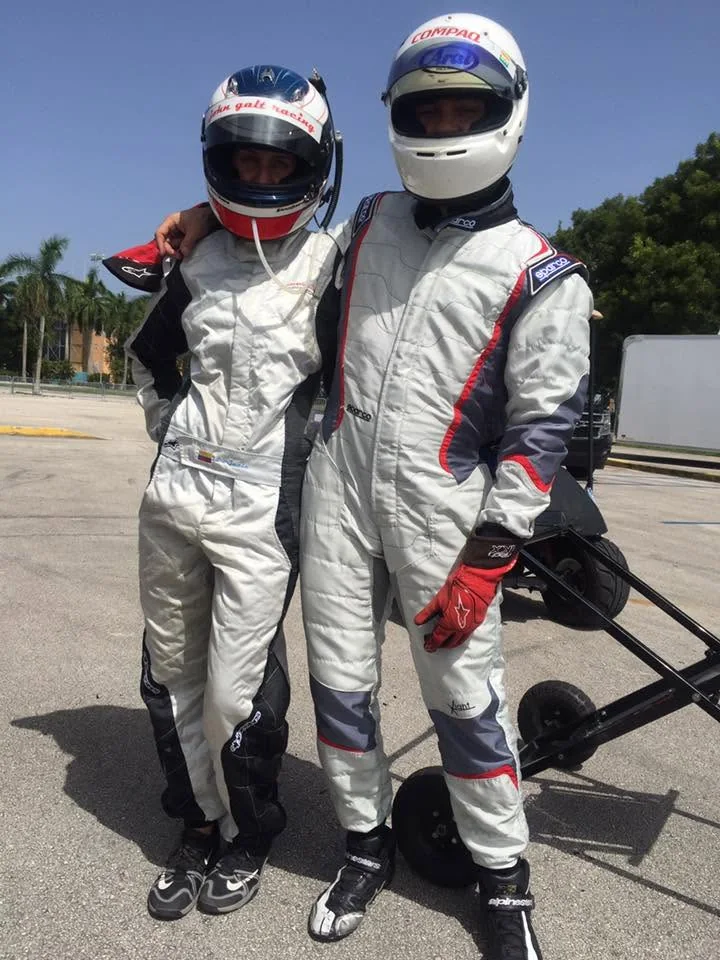 Two VMotorsport drivers in race suits and helmets beside a kart